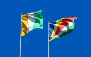 Ivory Coast and Seychelles flags waving on poles against a clear blue sky, symbolizing unity and pride.